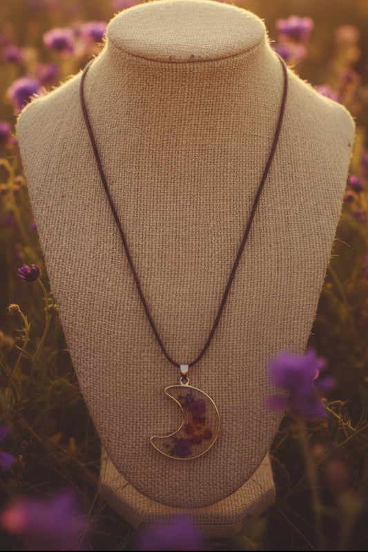 A crescent moon pendant necklace featuring dried purple and red flowers suspended from a brown cord.