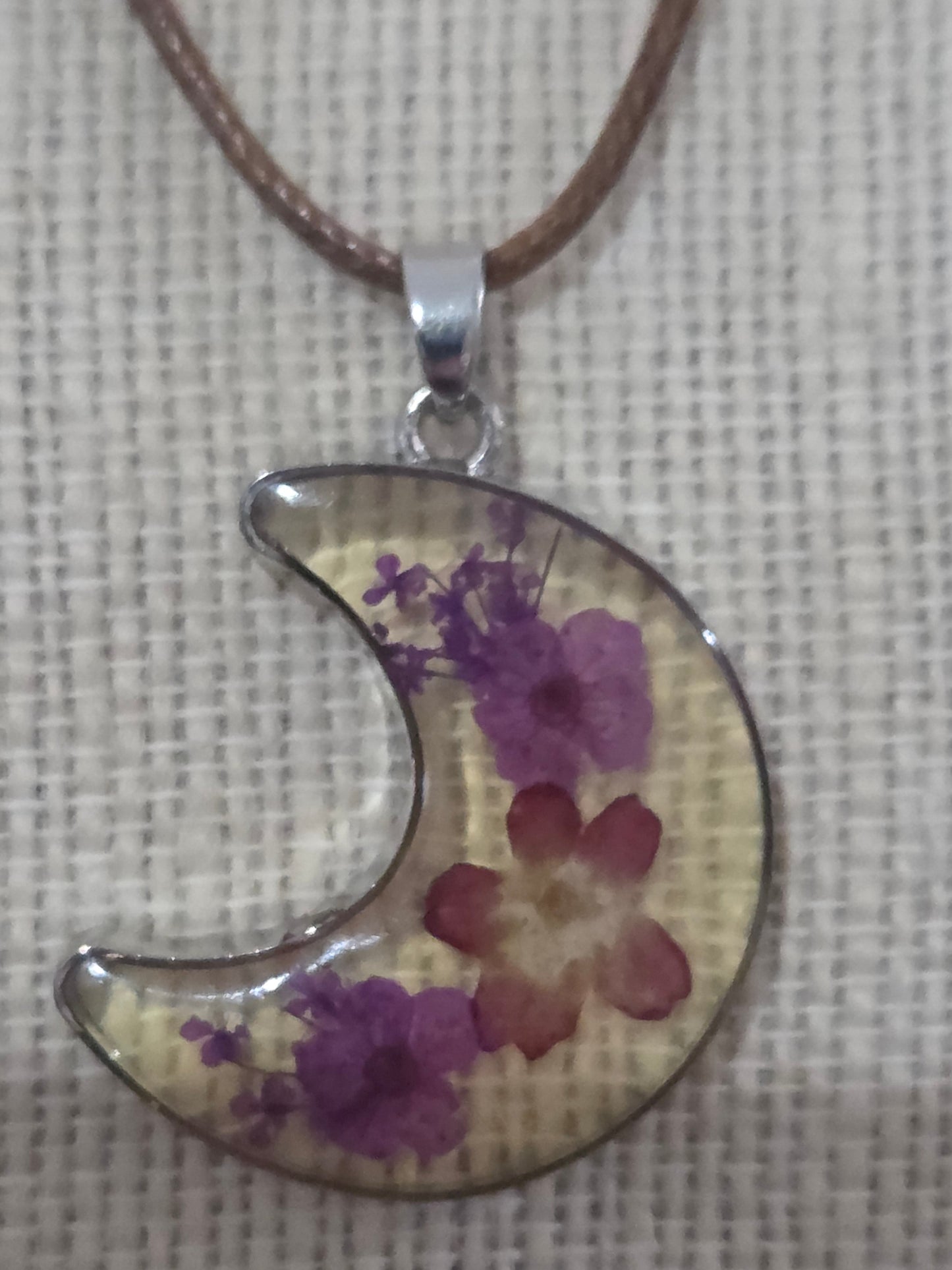 A crescent moon pendant necklace featuring dried purple and red flowers suspended from a brown cord.