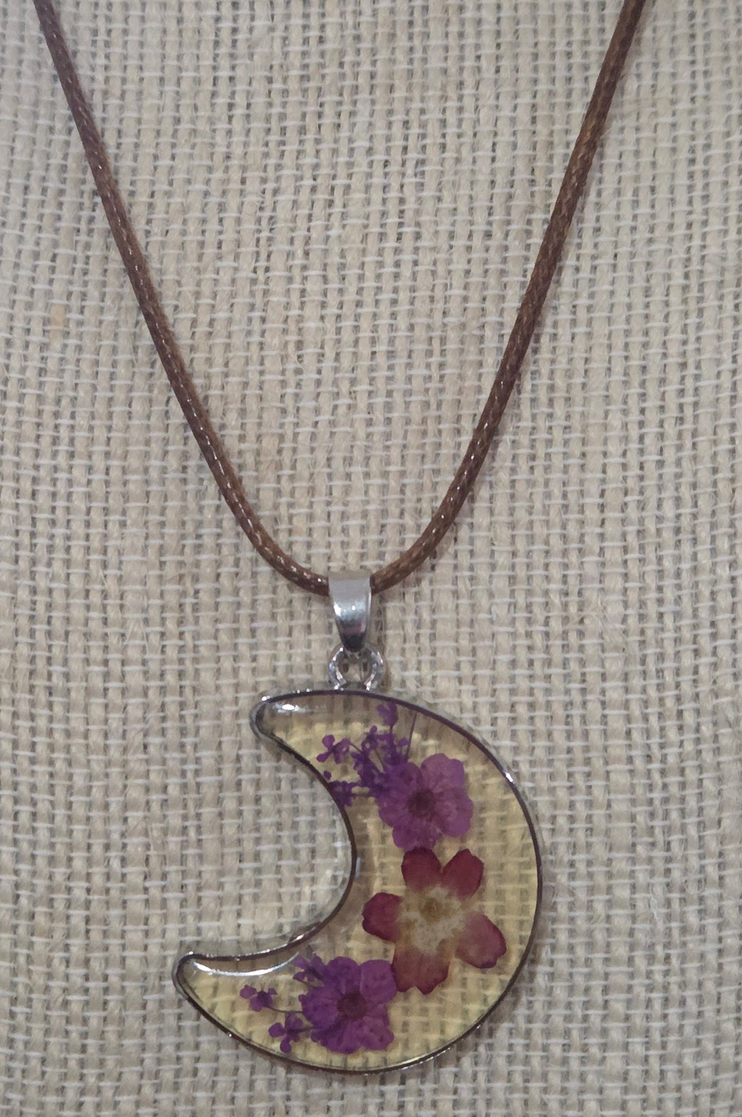 A crescent moon pendant necklace featuring dried purple and red flowers suspended from a brown cord.